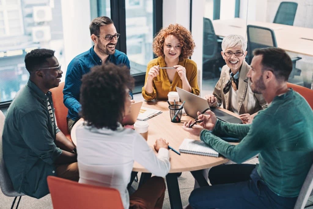 Six colleagues seated around a table in a bright office, talking over laptops and notes.