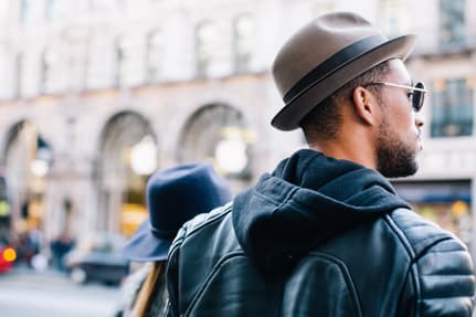 Person in a fedora, sunglasses, and black leather jacket, photographed from behind on a city street.